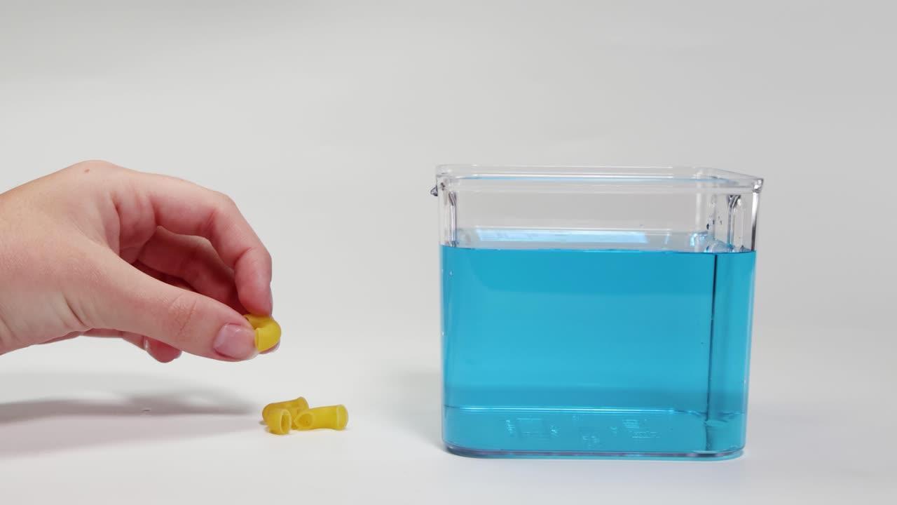 A hand drops pasta into a blue liquid, observing its buoyancy changes in a clear container