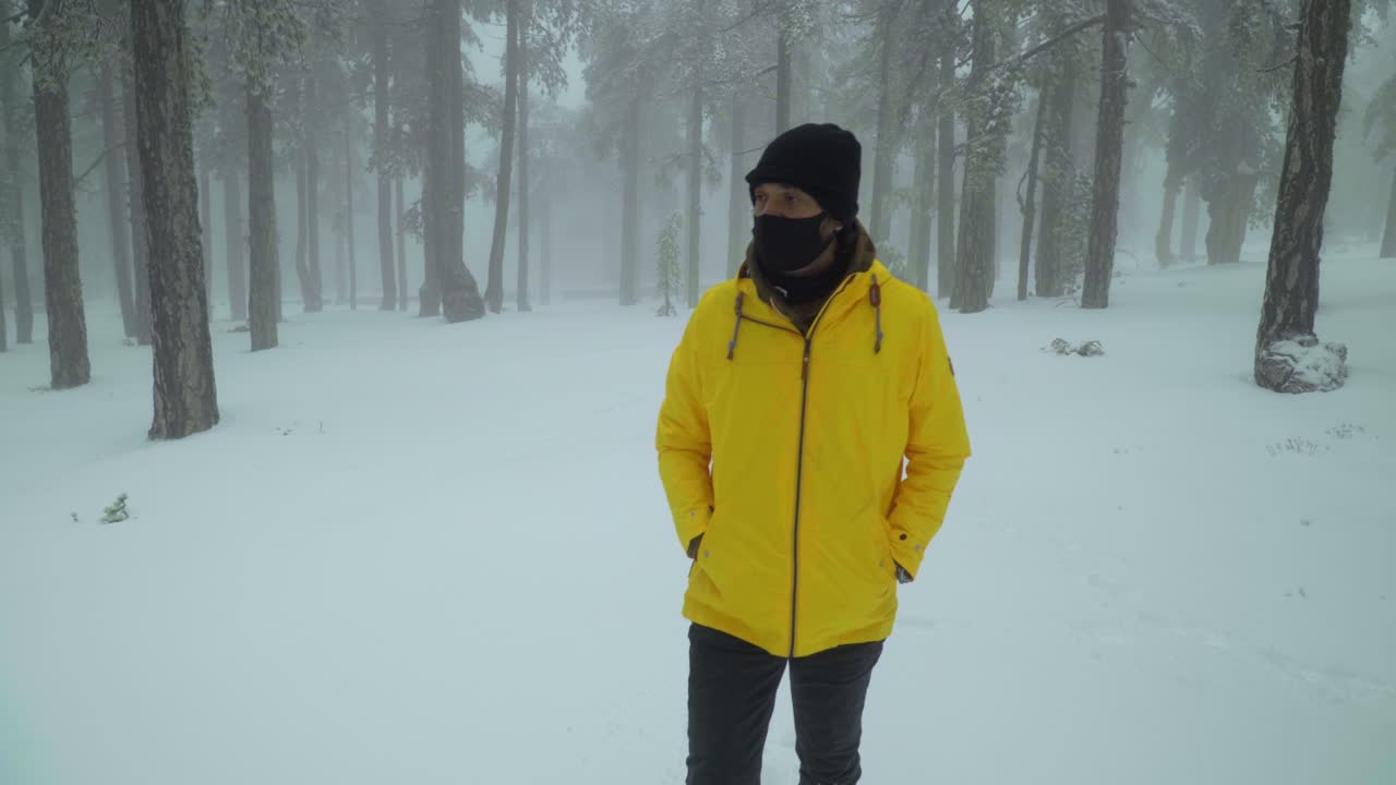 Front view following a man walking alone through a snowy forest, while wearing a bright yellow coat