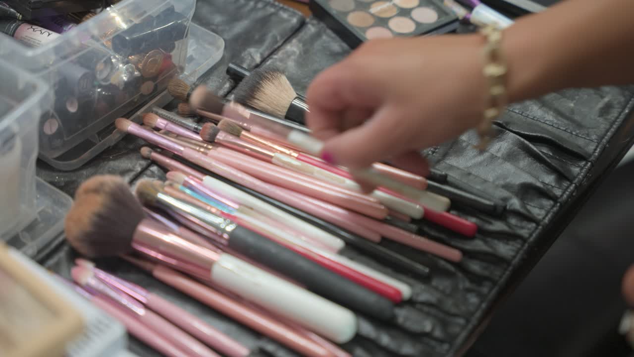 Un primo piano delle mani di una truccatrice che sceglie un pennello specifico da un set professionale. Il kit include vari pennelli, spugne e palette disposti per una sessione.