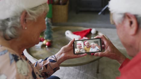 Pareja-Caucásica-Mayor-Usando-Un-Teléfono-Inteligente-Para-Una-Videollamada-Navideña-Con-Una-Familia-Feliz-En-La-Pantalla