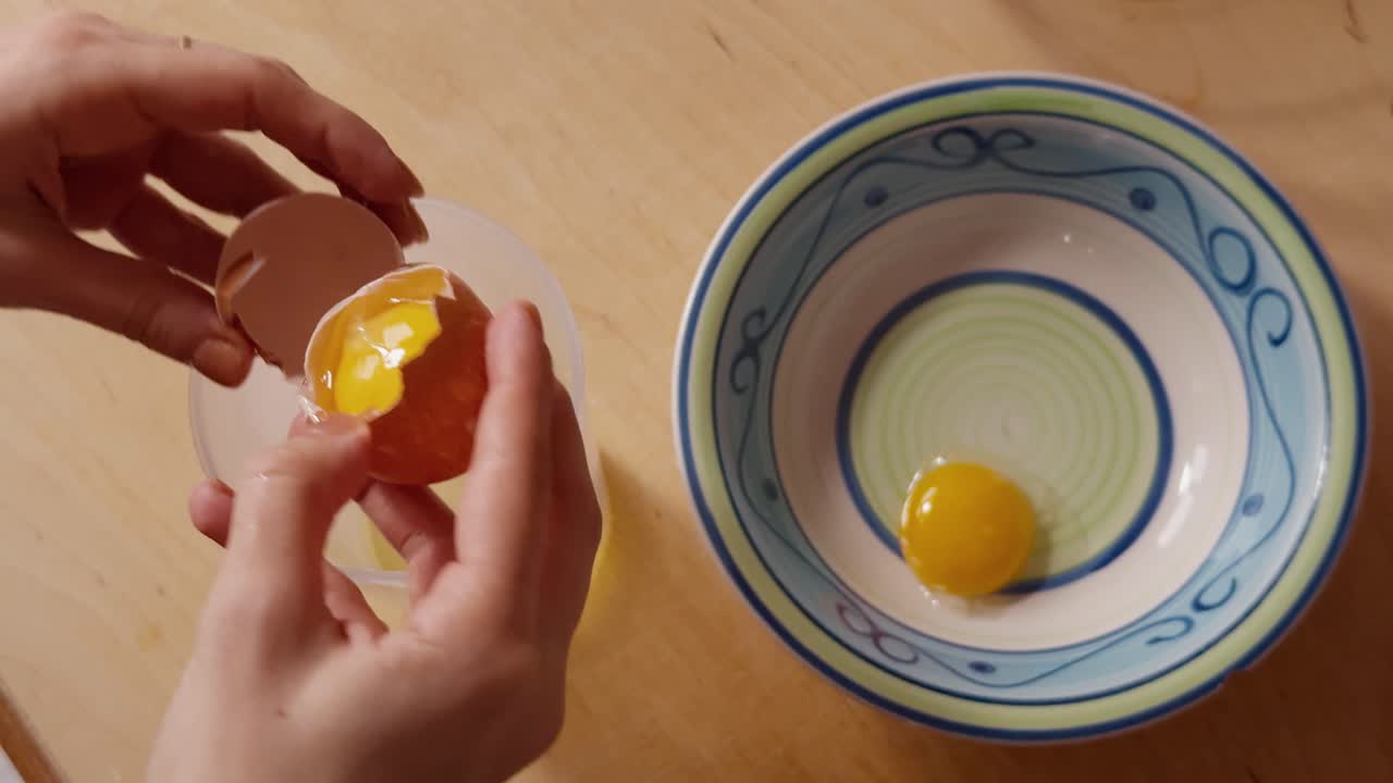 A person carefully separates an egg yolk from the white, holding the cracked shell above a bowl and container