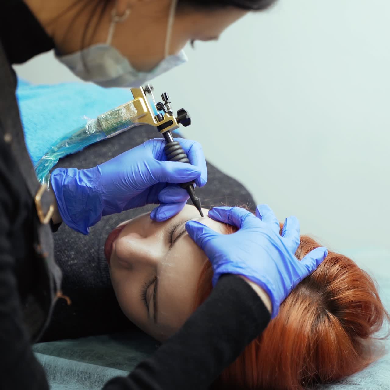 A woman in gloves is holding the client with one hand, and doing the eyebrow tattoo in the beauty salon with the other hand. Permanent makeup technique