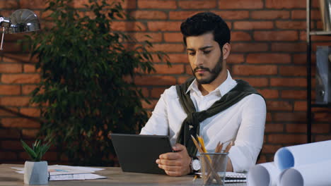 Young-Arabian-Architect-Sitting-At-Desk-In-His-Office-And-Working-With-Some-Documents-And-Tablet