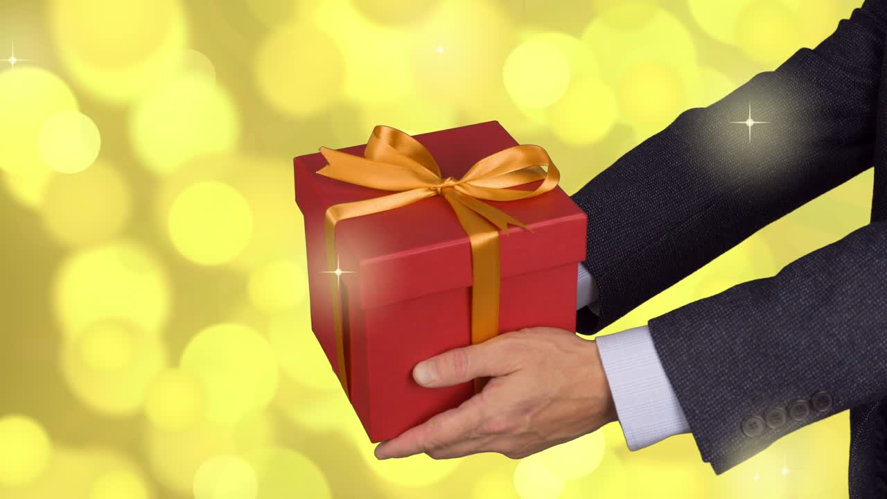 두 남자의 손은 황금 활과 함께 빨간 선물 상자를 들고 있습니다. 이브 선물을 축하합니다. 고전적인 슈트를 입은 카우카시안 남자. 황금 빛의 배경. 잠겼습니다.