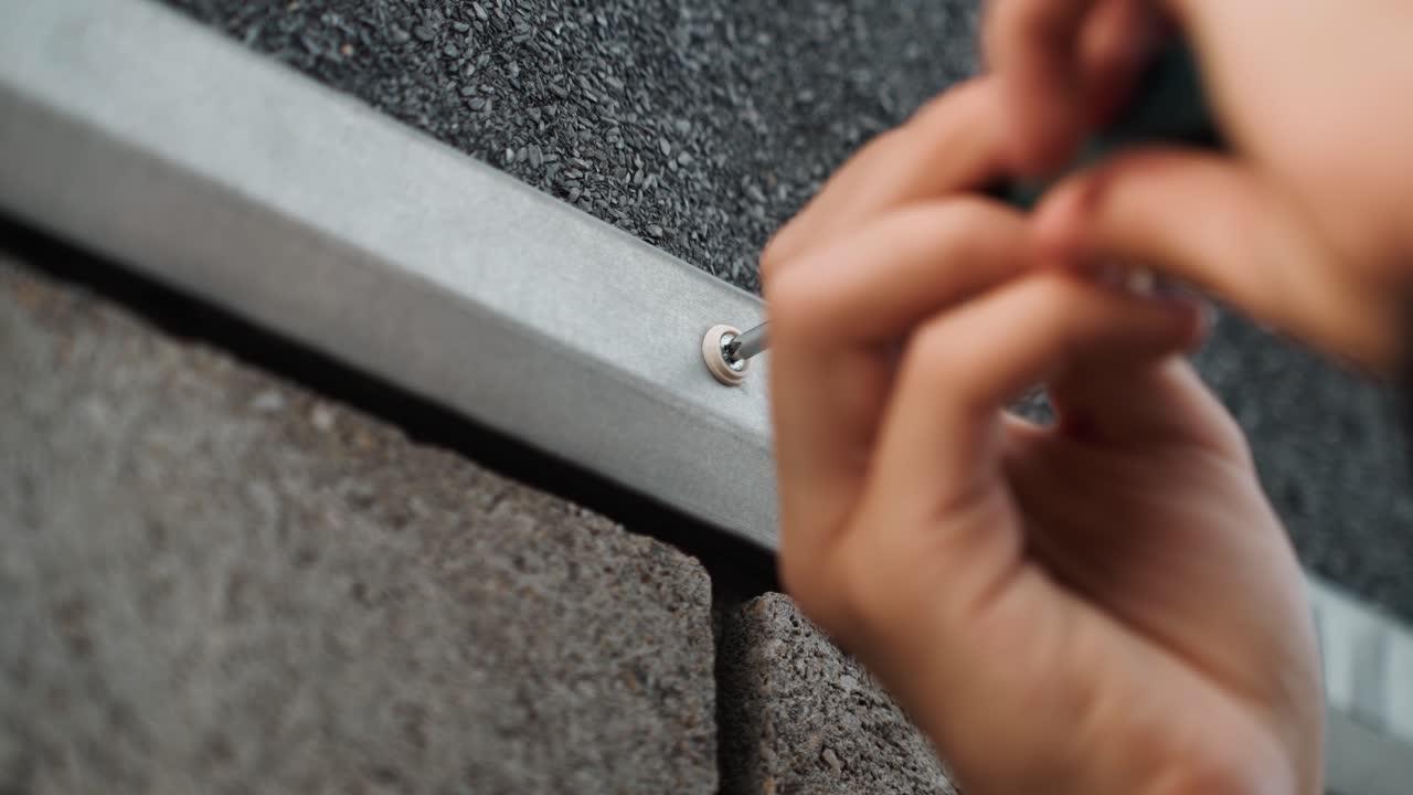Close-up de mãos apertando parafuso de rufo de telhado, faz-tudo usando chave de fenda para fixar rufos metálicos sob telha asfáltica em parede exterior, borda de concreto visível, profundidade de campo rasa e focada.