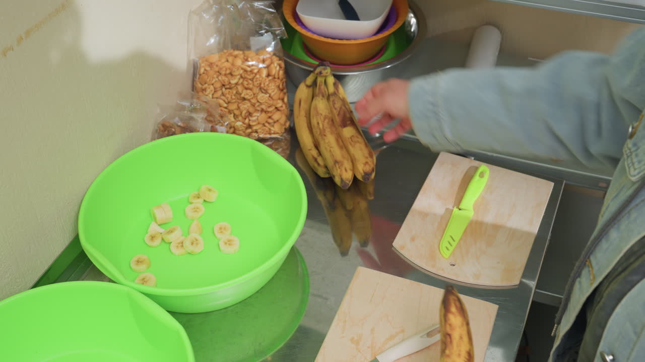 green bowls on stainless counter, bananas and cereal hands reach for bunch beside cutting board and knife, tidy pantry items and scale visible, cozy morning meal prep with organic fruit and simple