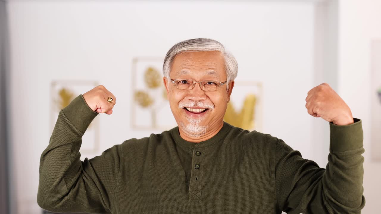 An elderly Asian man in a green shirt smiles and flexes his arms confidently in a well-lit, modern living room with soft natural lighting