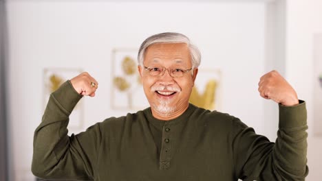 an elderly asian man in a green shirt smiles and flexes his arms confidently in a well-lit, modern living room with soft natural lighting