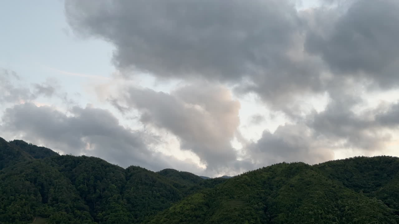 Fast-moving clouds cross the sky over a green mountain ridge during dusk. The pastel colors add a peaceful atmosphere.