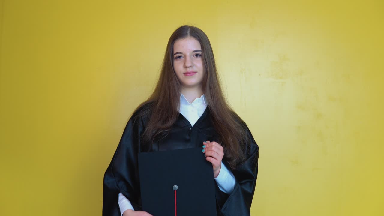 un graduado universitario está de pie frente a la cámara sosteniendo un sombrero de maestro con una borla roja. foto delantera de una mujer joven con el cabello largo y liso en un fondo amarillo