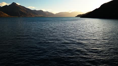 Plataforma-Rodante-Aérea-Sobre-El-Lago-Wakatipu-Mientras-La-Puesta-De-Sol-Brilla-Sobre-La-Ladera-Bajo-Un-Cielo-Azul