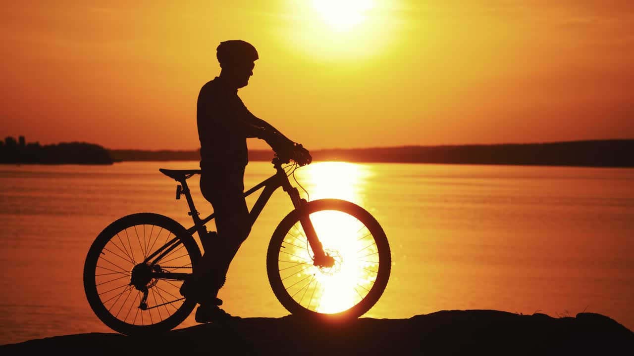 Silhouette of a cyclist near the river at sunset. Sporting man in helmet and glasses rides his bike on the background of beautiful sunset over the water surface.