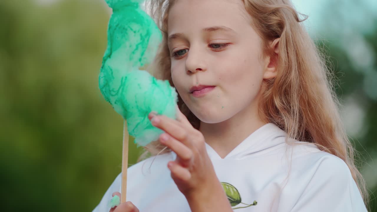 Portrait of a child eating green cotton candy. Little blonde girl is eating sweet candy-floss in the park.