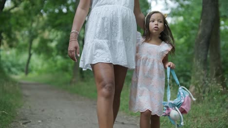 una mujer embarazada y su pequeña hija, ambas vestidas de blanco, caminan de la mano a través de un parque cubierto de hierba. la escena captura un momento sereno de unión familiar en la naturaleza, rodeada de árboles.
