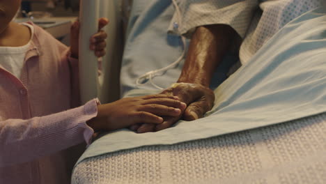 Niña-Tocando-La-Mano-Del-Abuelo-Acostado-En-La-Cama-Del-Hospital-Niño-Mostrando-Afecto-Al-Lado-De-La-Cama-Por-El-Abuelo-Recuperándose-De-Una-Enfermedad-Atención-Médica-Apoyo-Familiar