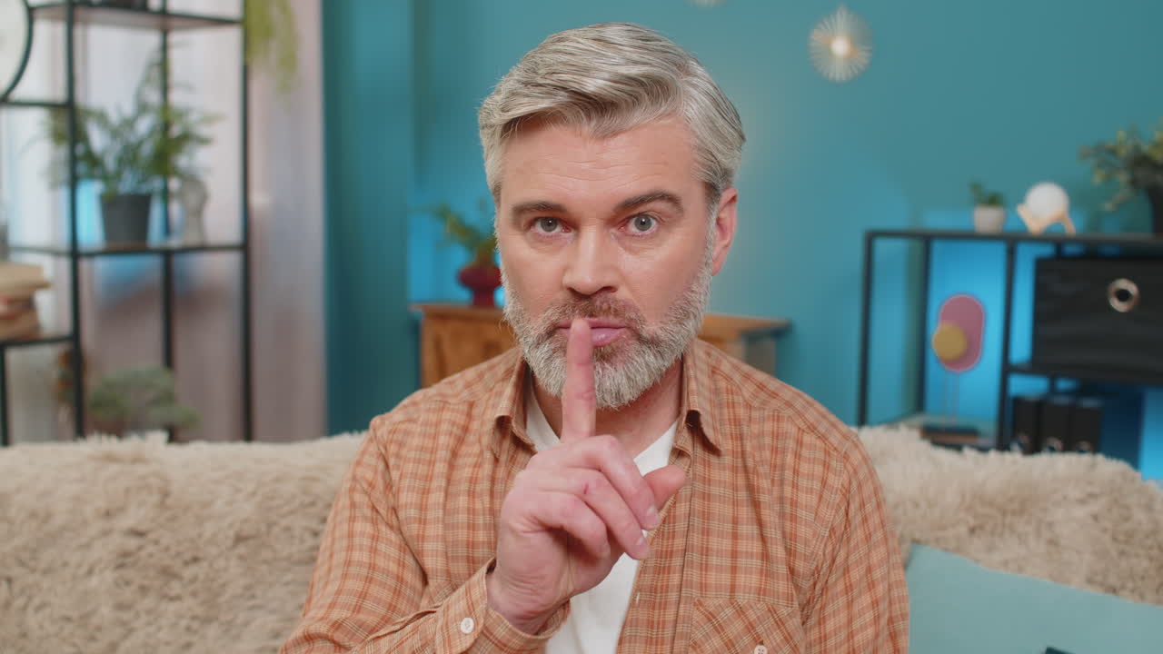 Mature man sitting on sofa making quiet gesture expressing seriousness and secrecy at home