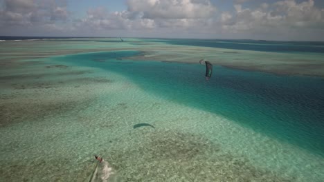 Un-Kitesurfista-Se-Desliza-Sobre-Aguas-Cristalinas-De-Color-Turquesa-Cerca-De-Una-Isla-Tropical,-Vista-Aérea