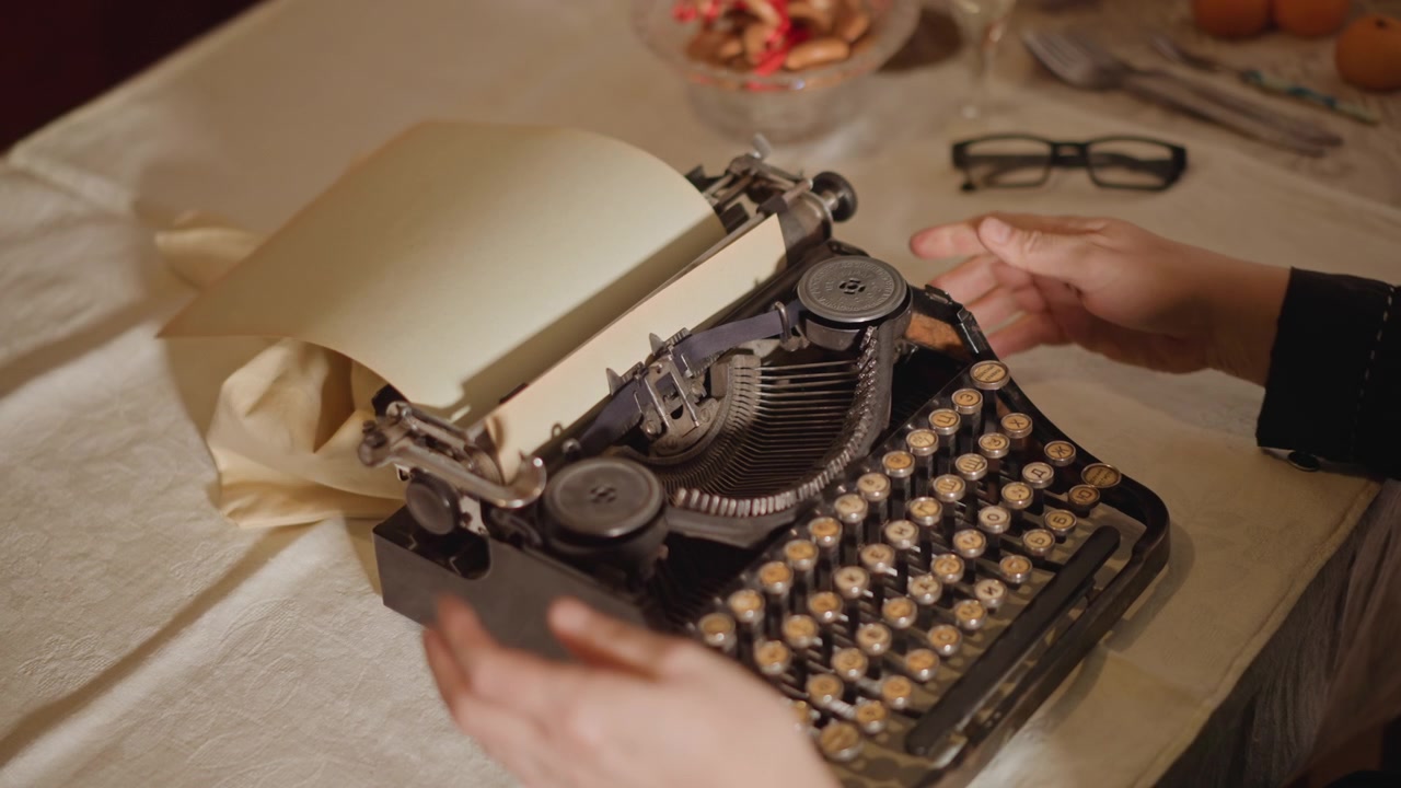 Hands loading blank paper into typewriter, cozy desk scene with linen tablecloth and glasses, author adjusting carriage and ribbon, warm amber light, closeup on mechanical keys and tactile ritual