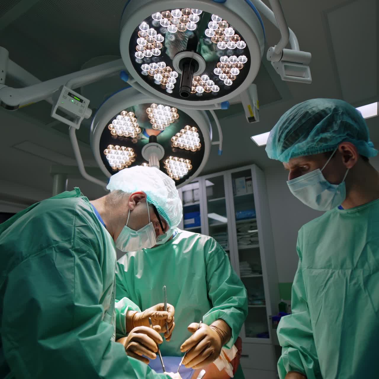 Team di professionisti della chirurgia che opera il paziente. I dottori utilizzano strumenti di metallo al lavoro. Due lampade rotonde illuminano la sala operatoria.