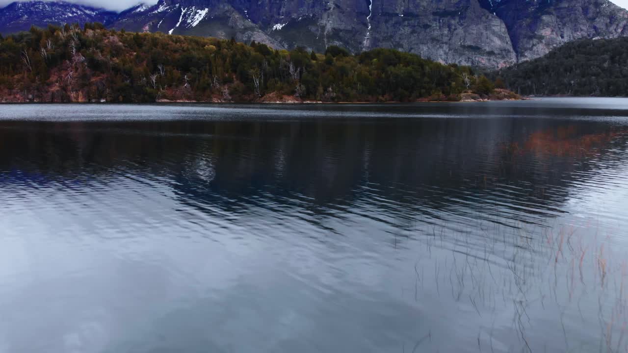 An aerial perspective, gliding over a lake with reflective waters, unveiling the grandeur of Patagonia's snow-dusted mountain peaks and their verdant forested slopes