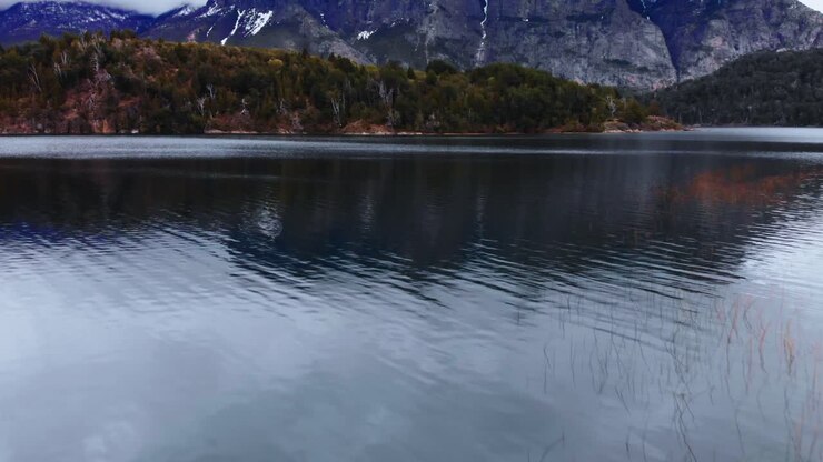 An aerial perspective, gliding over a lake with reflective waters, unveiling the grandeur of Patagonia's snow-dusted mountain peaks and their verdant forested slopes