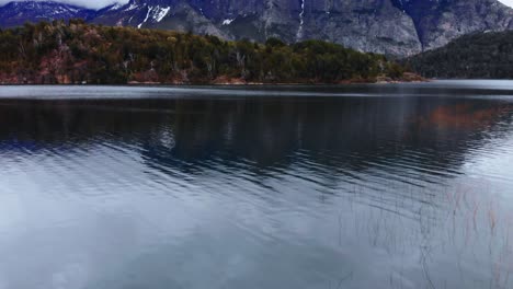 Una-Perspectiva-Aérea,-Deslizándose-Sobre-Un-Lago-Con-Aguas-Reflectantes,-Revelando-La-Grandeza-De-Los-Picos-Nevados-De-La-Patagonia-Y-Sus-Verdes-Laderas-Boscosas