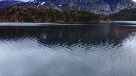 an aerial perspective, gliding over a lake with reflective waters, unveiling the grandeur of patagonia's snow-dusted mountain peaks and their verdant forested slopes