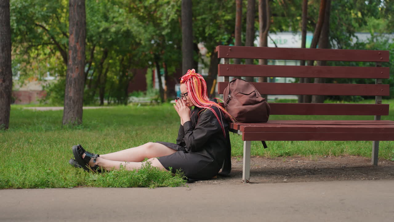 공원 잔디에 앉아 있는 핑크색 머리 소녀, 공원 벤치 옆에 앉아 있는 십 대 백인 여성, 밝은 핑크색 드레드록을 한 젊은 여성이 공원에서 길거리 공연 중 휴식을 취하고 있어요.