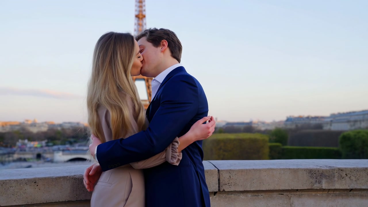 Young standing on a stone terrace, facing each other in an intimate pose as the woman rests her hands on the man’s arms. The man wears a dark suit