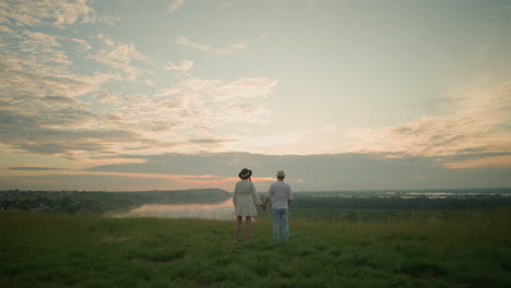 a couple stands on a grassy hill beside a serene lake at sunset, gazing at the sky. the man, in a white shirt, hat, and jeans, gently holds the woman hand she wears a black hat and a white dress