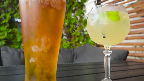 beer glass filled with beer that is foaming and bubbling in slow motion side by side with a cocktail glass filled with drink, ice and lime wedge. drinks are in sharp focus, background is blurred.