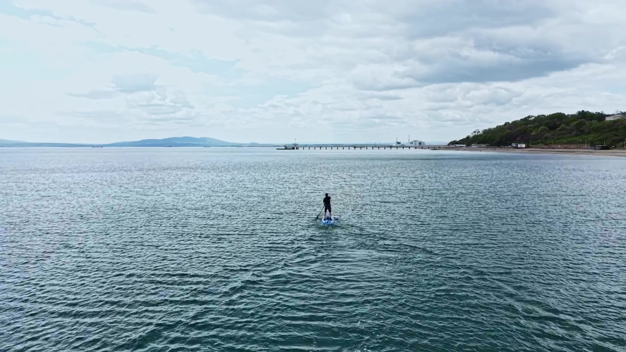 Les vagues éclaboussent autour d'une personne debout sur un paddleboard dans une grande étendue d'eau près d'une jetée au loin par une journée nuageuse
