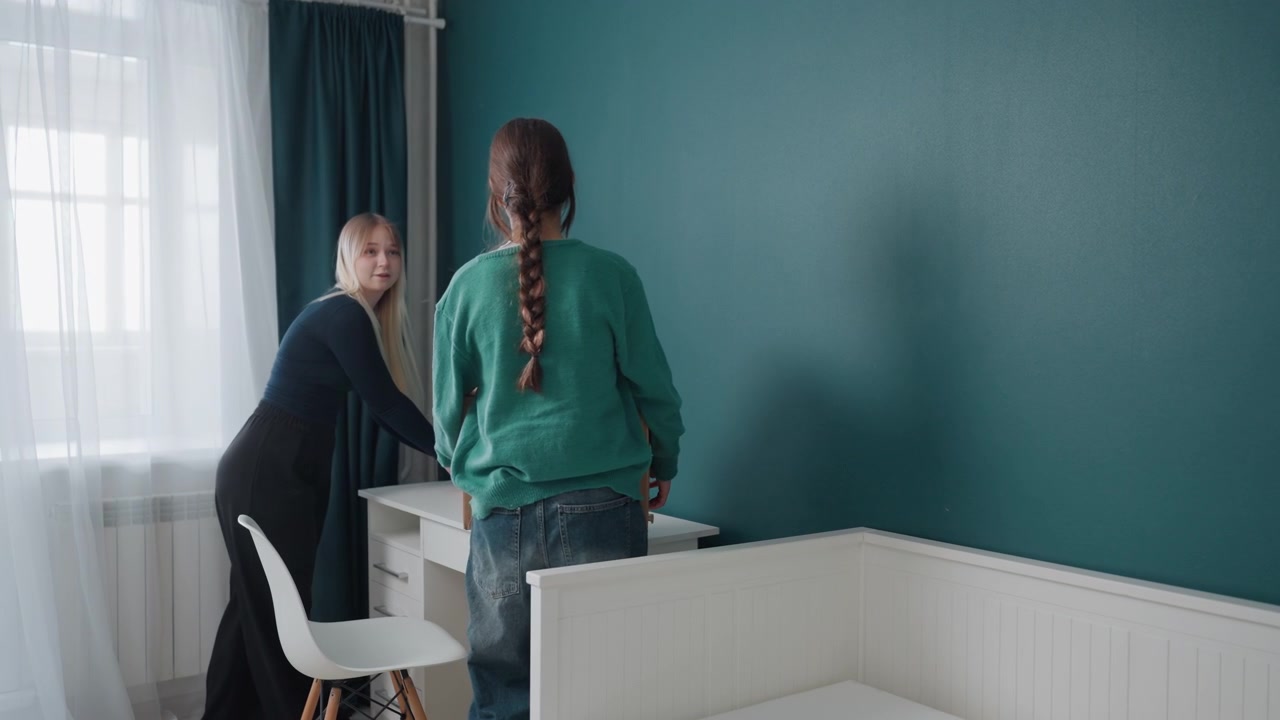 Young white roommates plan apartment layout near window and radiator, stacking boxes, testing chair placement, discussing decor and measurements, collaborative mood with sunlight through curtains