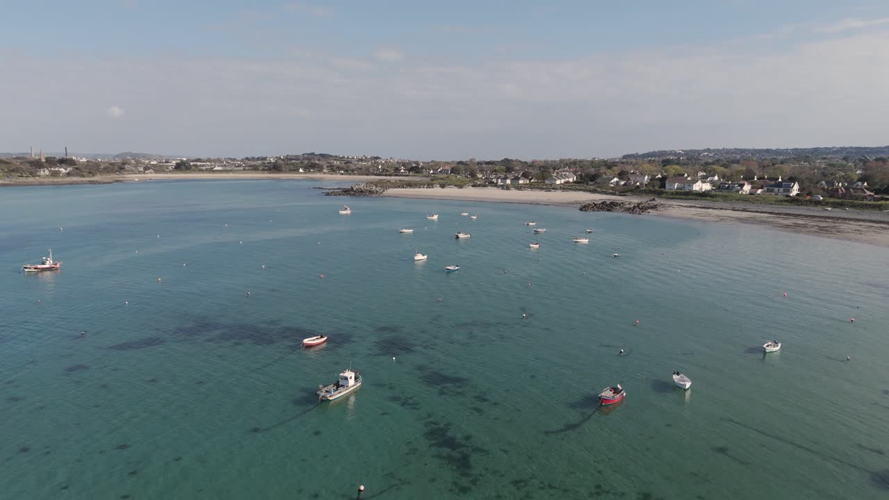 Vol de drone au-dessus d'une baie à Guernesey avec des bateaux au mouillage, une eau cristalline et des plages dorées par une journée ensoleillée et calme, vers les plages et les cottages en bord de mer