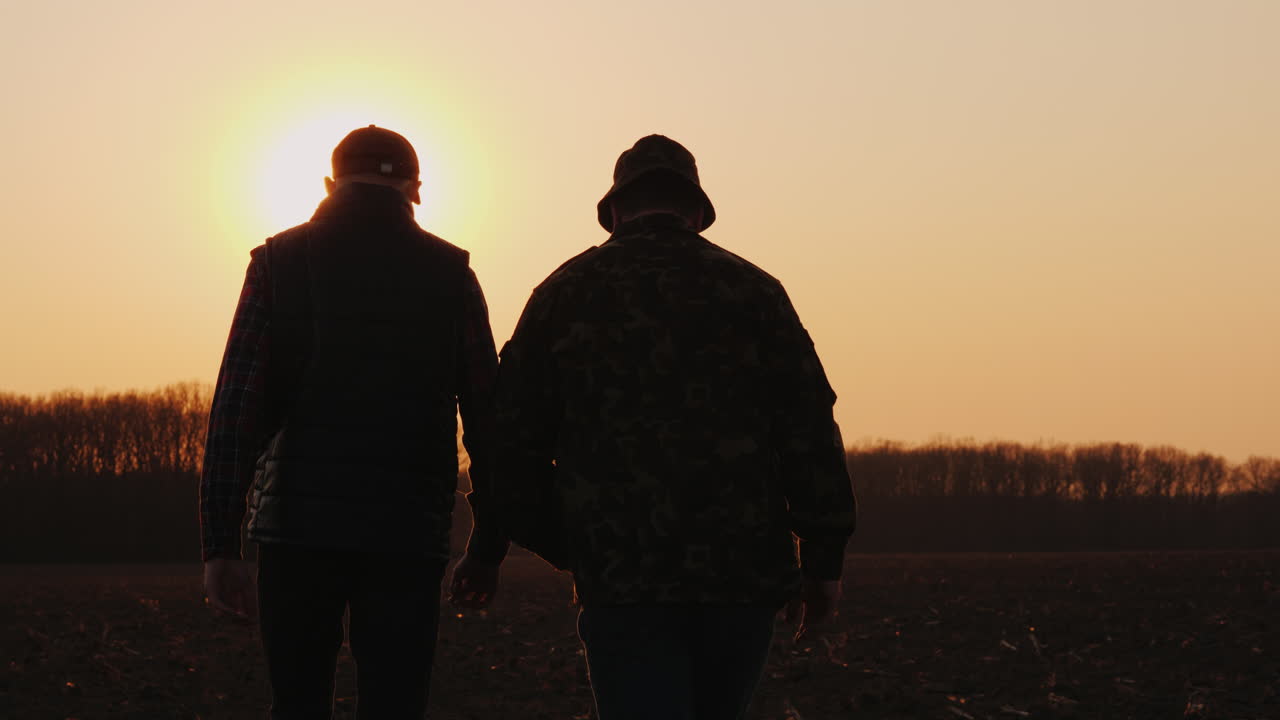 un anciano y un joven agricultor van juntos por un campo arado al atardecer