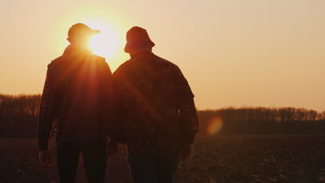 Ein-älterer-Und-Ein-Junger-Bauer-Gehen-Bei-Sonnenuntergang-Zusammen-über-Ein-Gepflügtes-Feld