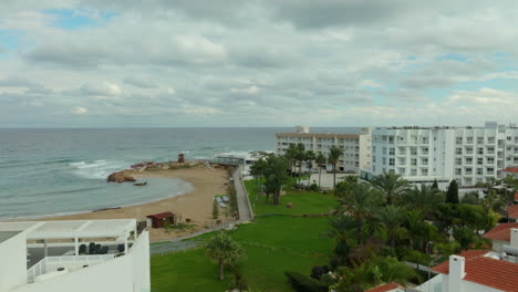 Overlooking-the-tranquil-seaside-of-Pernera-in-Cyprus,-small-sandy-bay,-gentle-surf,-and-a-waterfront-promenade-lined-with-palm-trees,-a-backdrop-of-modern-resort-buildings-under-a-cloudy-sky