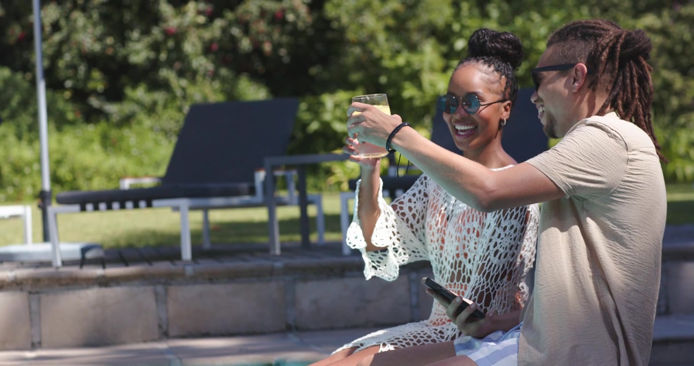 African American couple in swimsuits sharing as man tilting phone, passing and toasting drinks pool