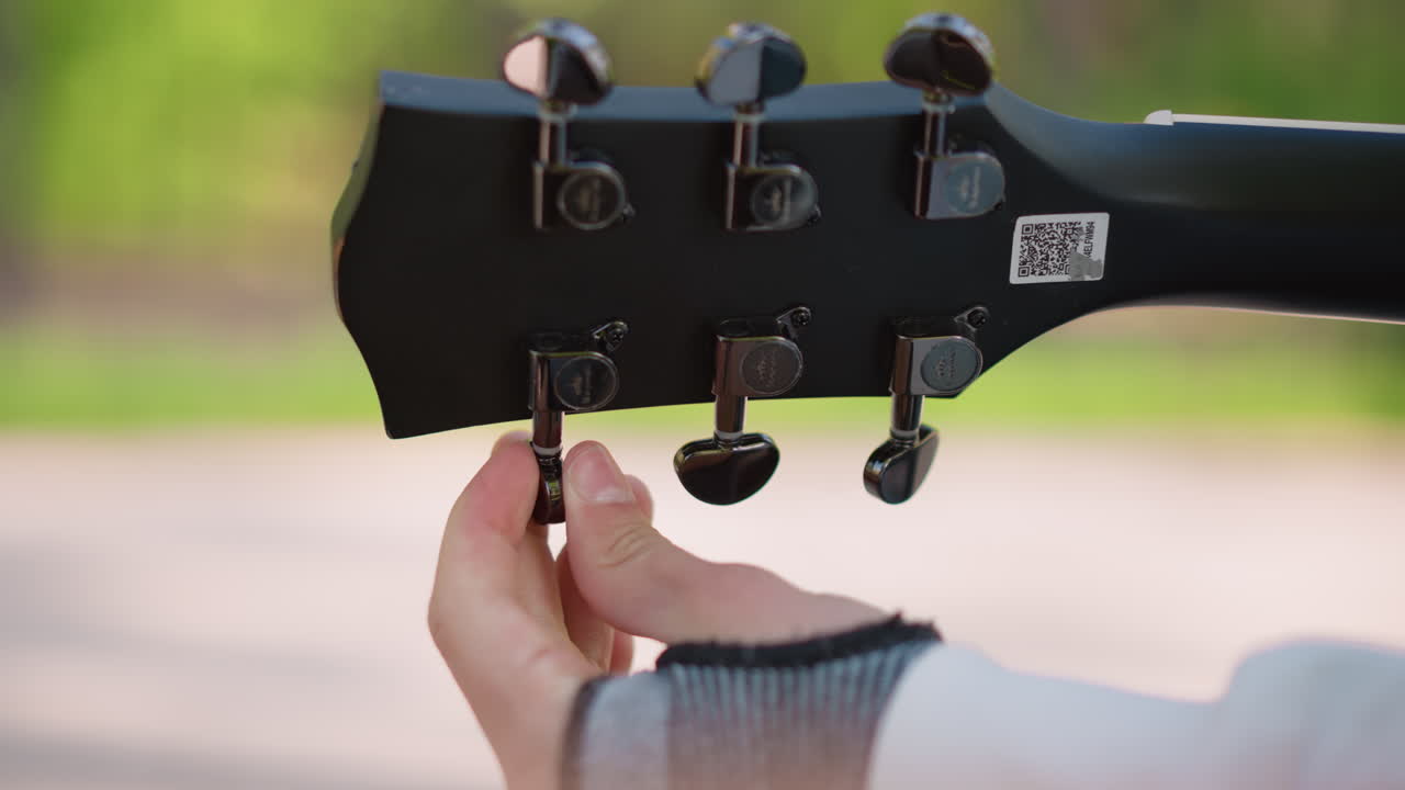 closeup hand tuning guitar headstock with fingerless glove in park, musician adjusts chrome tuning pegs on black acoustic neck, warm sunlight and soft bokeh create calm, intimate preparation