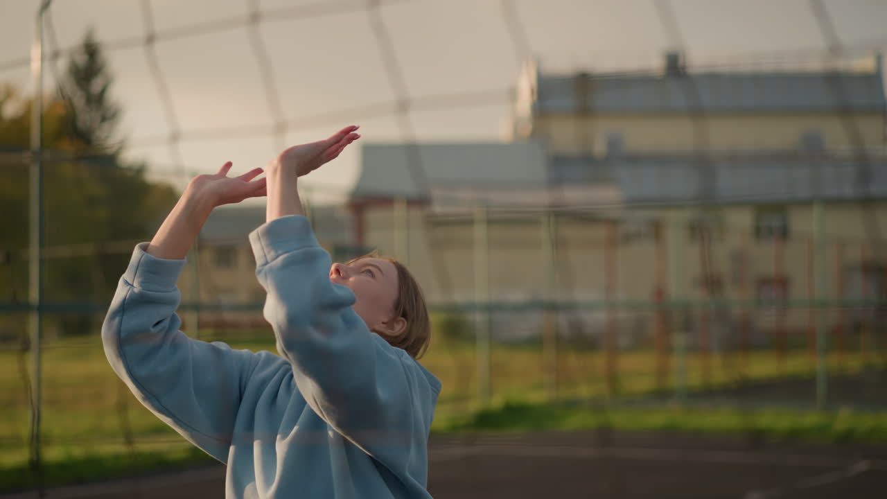 joven jugando al voleibol al aire libre, ajustando su posición cerca de la red de voleibol, con un fondo borroso con vegetación y edificios en la distancia, iluminados por la luz del sol