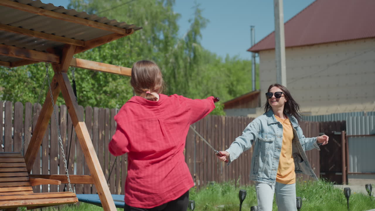 Two cheerful women dressed in casual outfits engage in playful sword duel beside wooden swing in sunny backyard with fence, green grass, and clear sky, sharing laughter