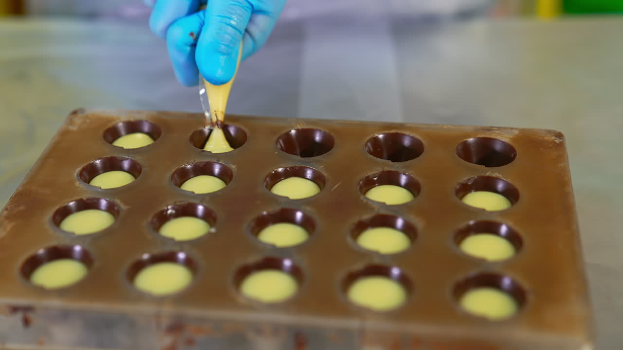 Candy molds covered with chocolate are being stuffed with creamy filling. Chocolatier using pastry bag for filling sweets. Close up.