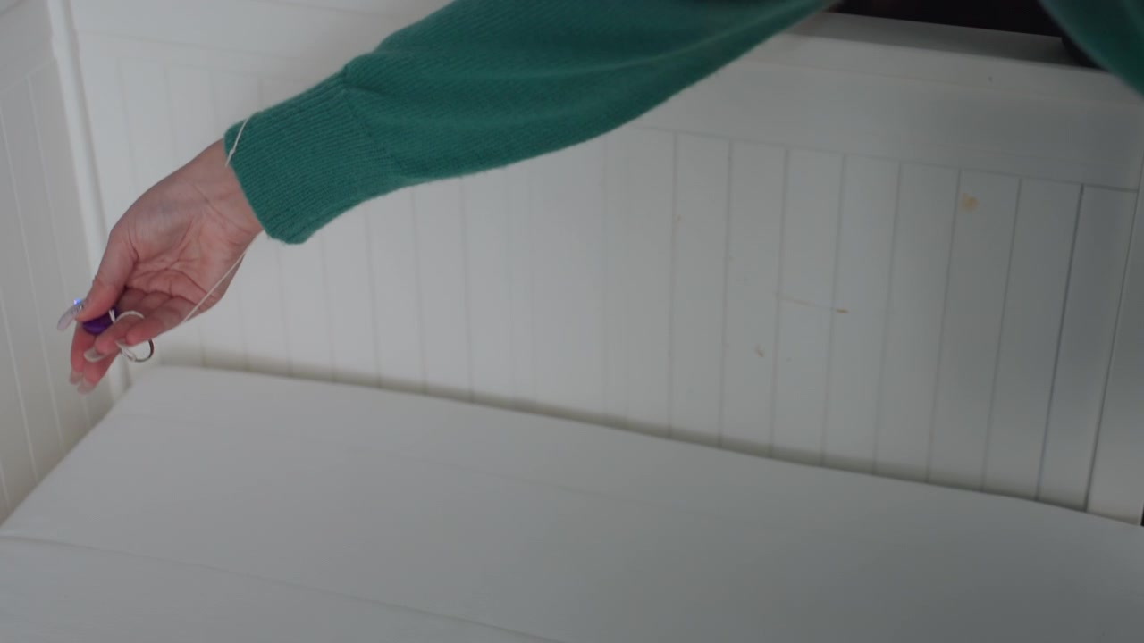 Donna che controlla la cucitura del materasso con primi piani delle mani illuminati, usando una piccola torcia portachiavi vicino all'angolo mentre tiene un vaso di piante. Ispezione metodica di trasloco dell'affittuario per macchie e residui, con attenzione e cautela.