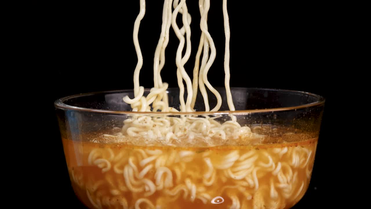 A hand skillfully lifts cooked instant noodles from a glass bowl of broth using chopsticks, set against a black background with bright, even lighting and a static camera