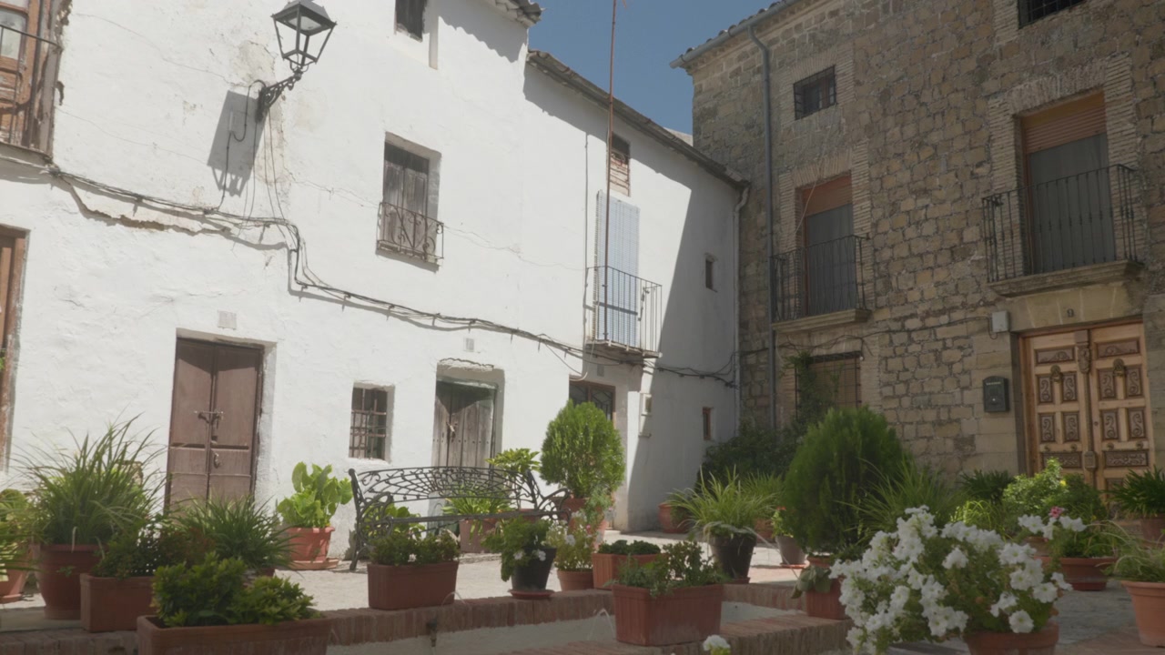 Un'inquadratura ampia e statica di un angolo tranquillo di un cortile a Iznatoraf, Jaén, Spagna. La scena presenta una combinazione di facciate rustiche imbiancate a calce e storici muri in pietra squadrata, adornati con vasi di fiori.