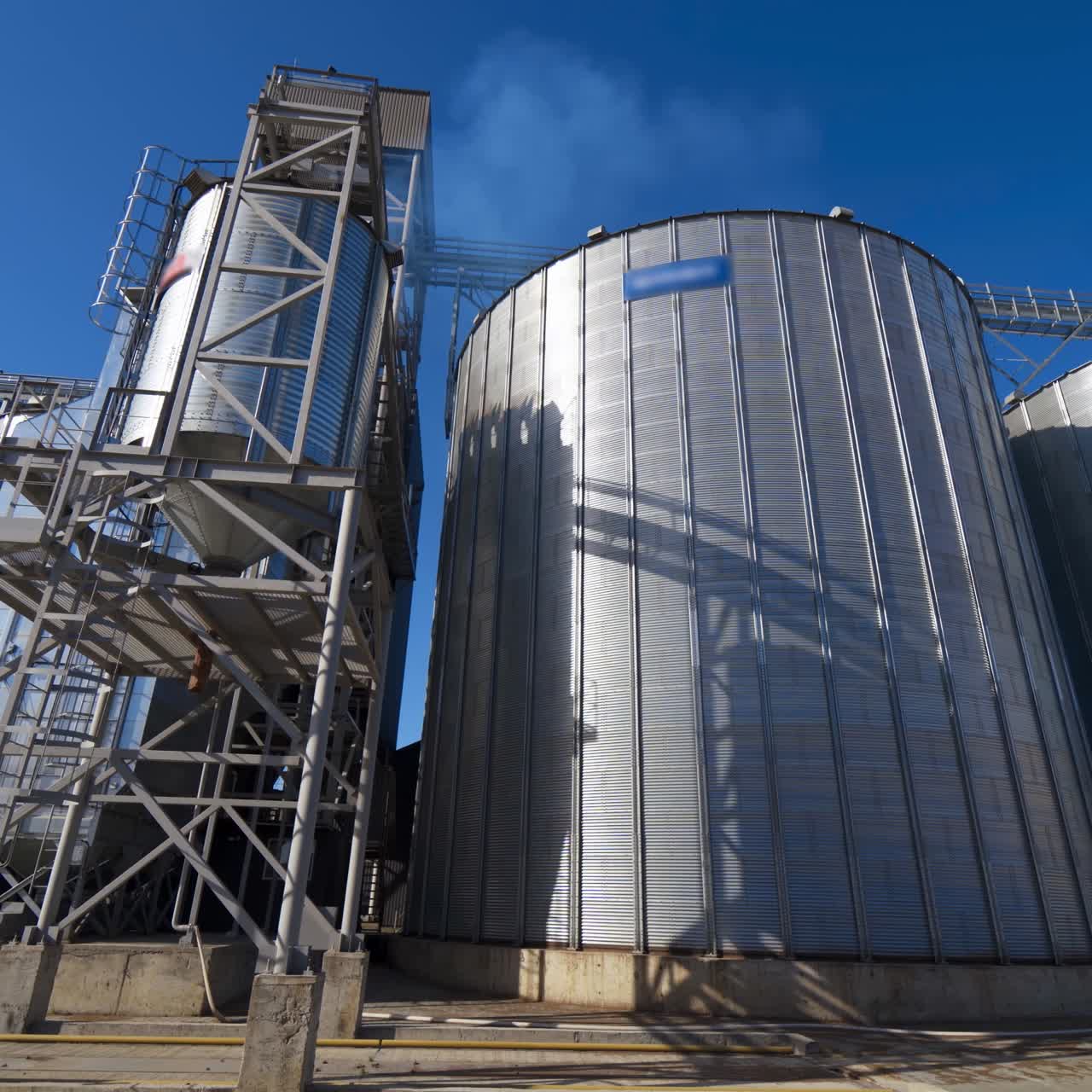 Modern factory for storage crop. Metal grain elevators on agricultural complex. Exterior of industrial plant under blue sky