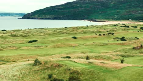 Aerial-drone-fly-over-rolling-hills-of-coastal-Irish-links-golf-course,-lush-greenery-and-ocean-backdrop,-establishment-shot