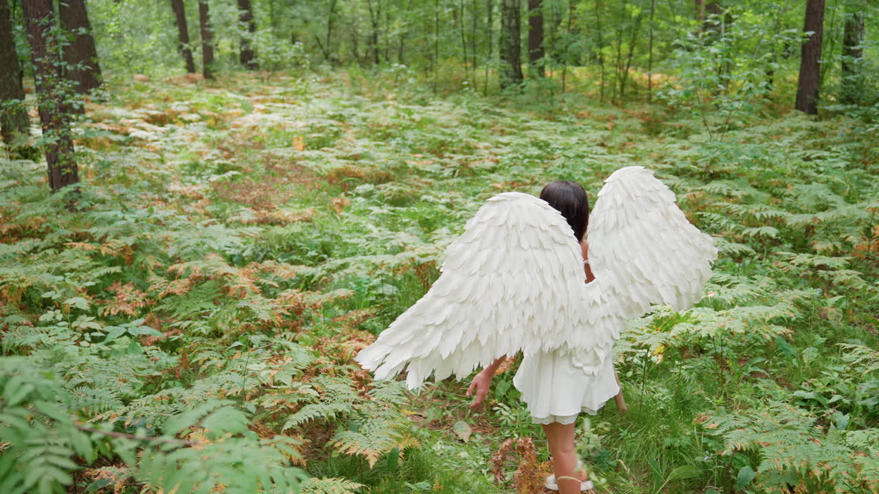 Girl dressed like divine figure with feathered white wings and flowing white dress walks through lush forest holding ancient wooden staff, embodying spiritual presence and mythical goddess energy