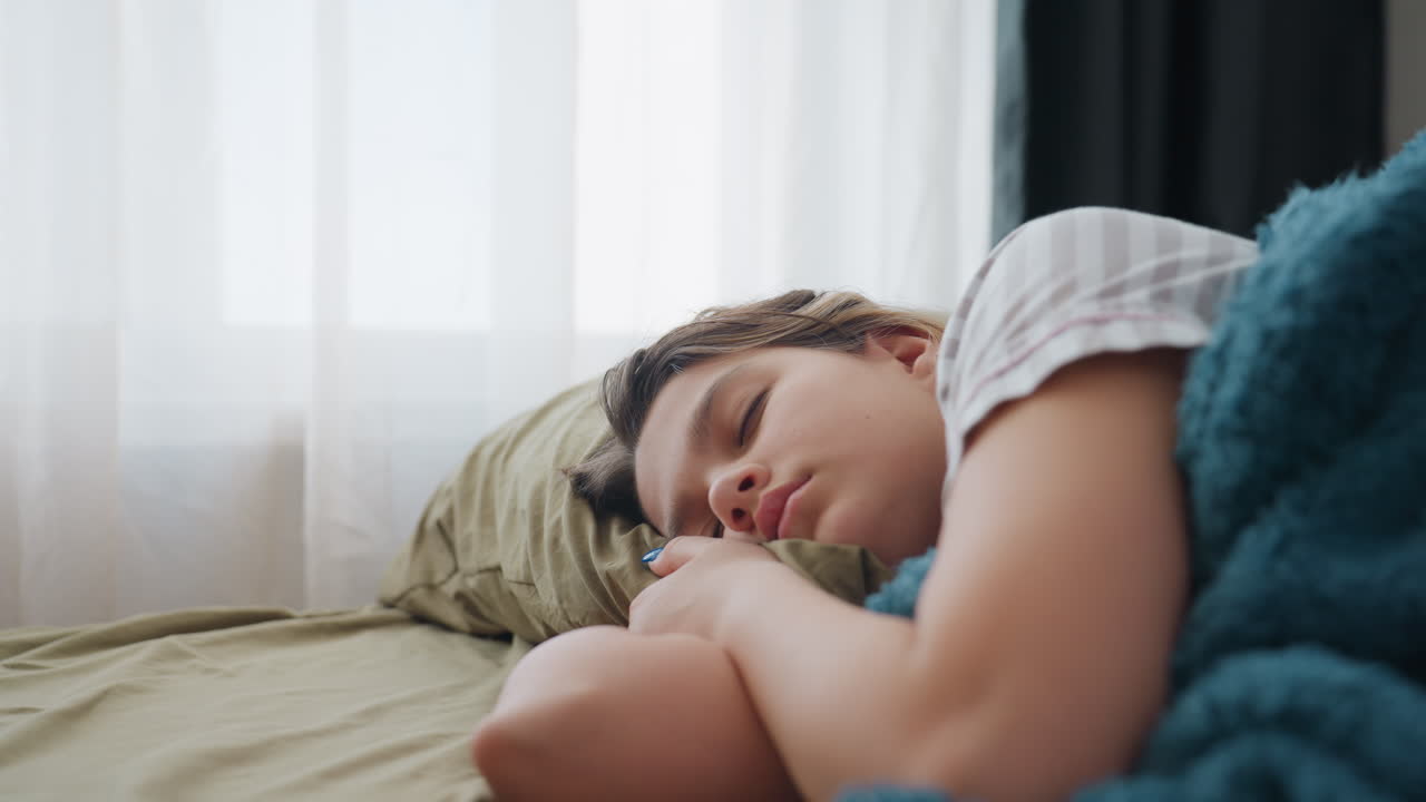 Young Woman Relaxes On Couch Under Blanket, Student Lying Peacefully On Sofa With Teal Blanket Nearby, Relaxed Female Student Taking Nap On Couch In Comfortable Domestic Environment
