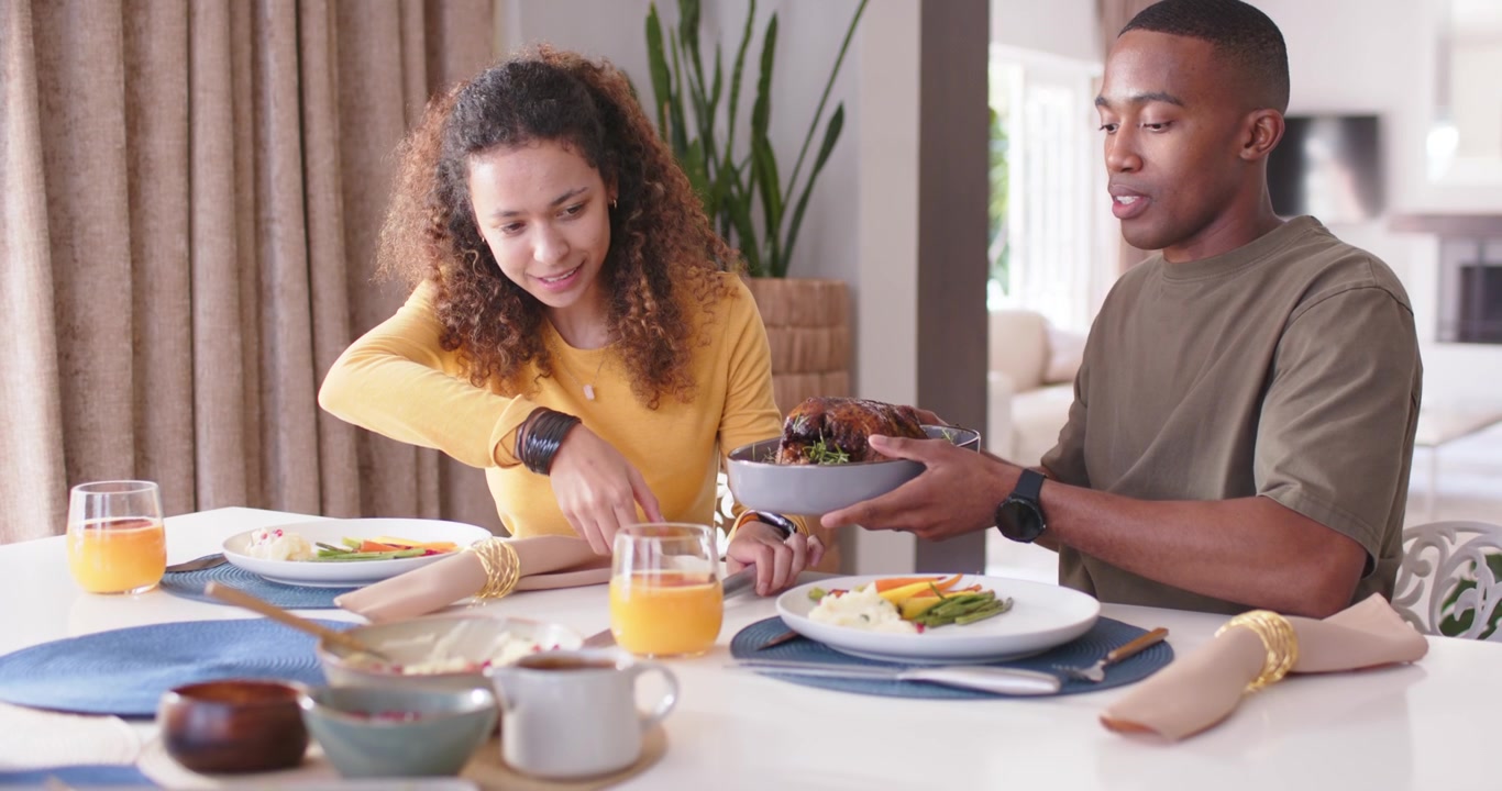 Couple at dining table, man bringing roast bowl, woman carving roast with knife to serve meal